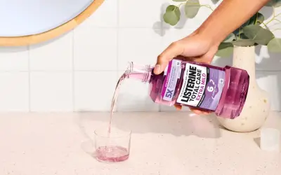 Ręka wylewa fioletowy płyn Listerine Total Care Extra Mild do szklanki. Poznaj listerine skład i ciesz się świeżym oddechem.